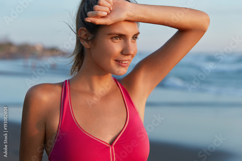 Outdoor shot of tired female athlete wears sports clothes, being sweaty, going to swim in sea, looks tired but satisfied with cardio training on coastline. Fatigue jogger wins sport competition
