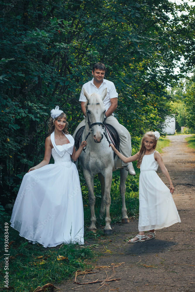 Man, woman dressed as a bride, girl and white horse in the park among the green trees