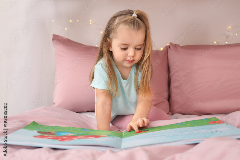 Cute little girl reading book while sitting on bed at home Cute little girl reading book while sitting on bed at home