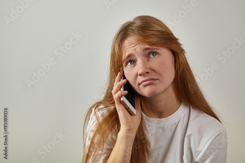 Headshot of young red-headed girl with freckles holding her phone next to her ear and talking with sad expression on her face