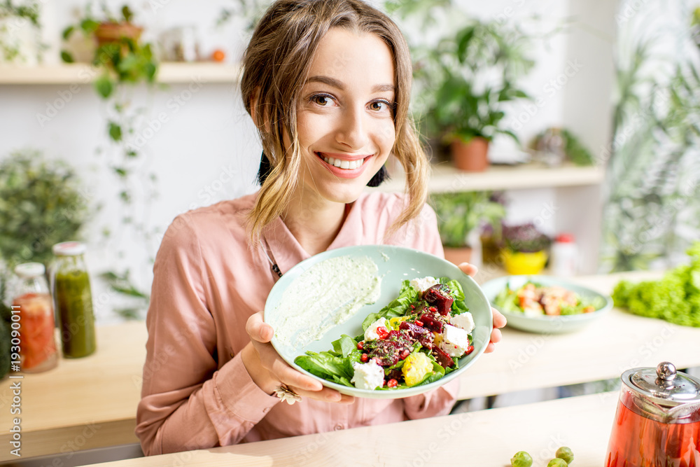 Plakat Portret młoda kobieta trzyma talerza sałatkowego obsiadanie indoors otaczający zielonymi kwiatami i zdrowym weganinem