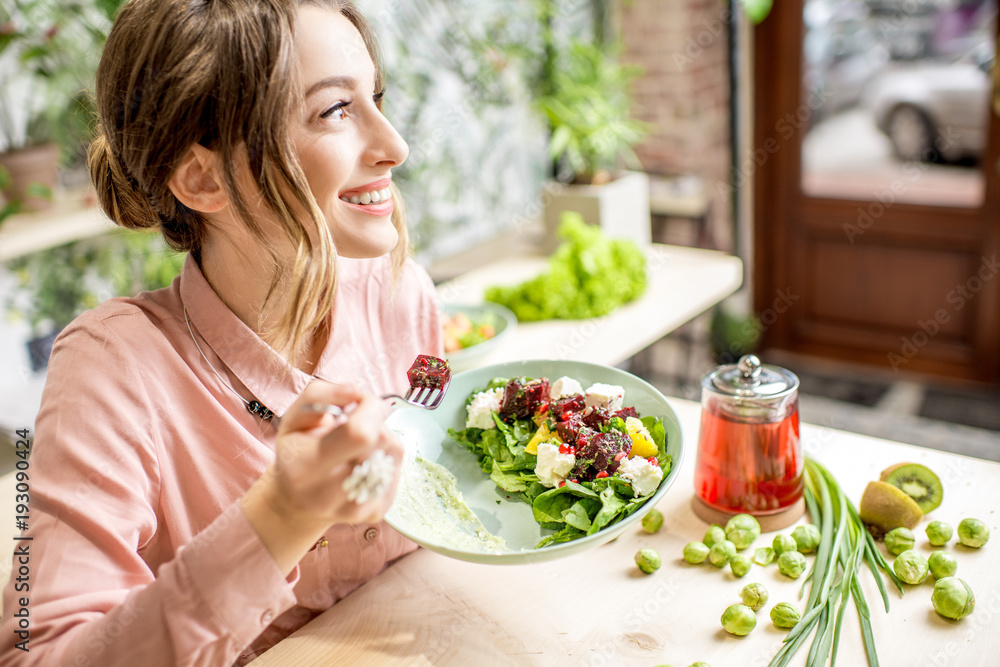 Young woman eating healthy food sitting in the beautiful interior with green flowers on the background