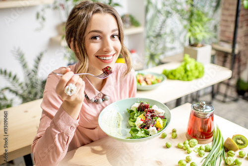 Leinwand Poster Young woman eating healthy food sitting in the beautiful interior with green flo