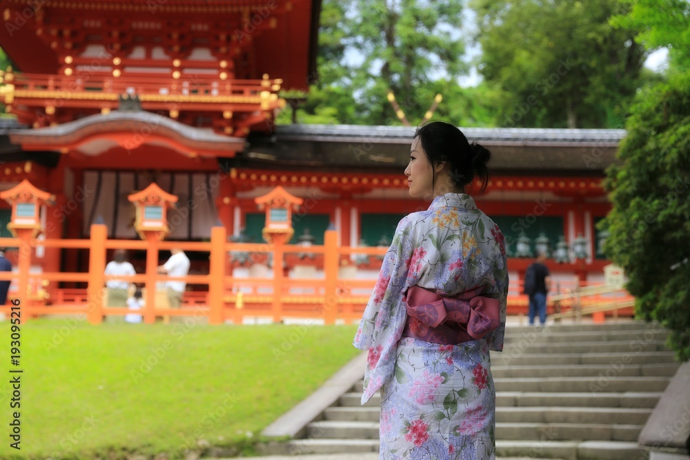 a young Japanese woman in summer kimono (yukata) standing in front of a red traditional Japanese building