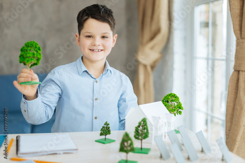 Involved in project. Upbeat pre-teen boy sitting at the table full of different miniature models and posing for the camera with a big smile while holding a tree model