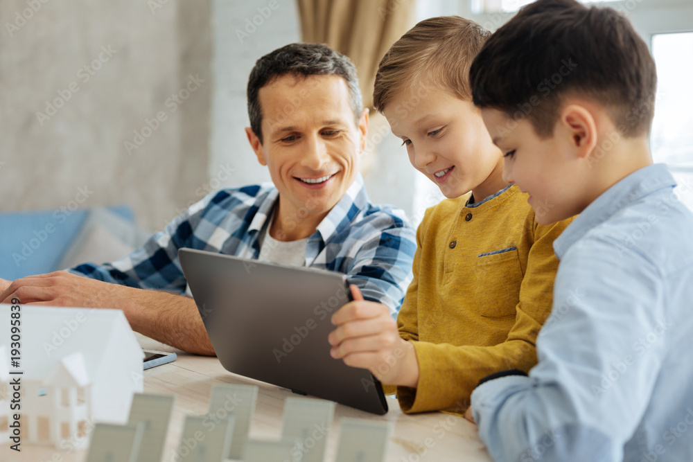 Quality time. Pleasant young man taking a break from work and devoting some time to his lovely sons, watching a video on the tablet with them Quality time. Pleasant young man taking a break from work and devoting some time to his lovely sons, watching a video on the tablet with them