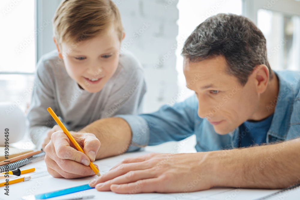 Fascinated by work. Pleasant curious pre-teen boy sitting at the table next to his father and watching him draw a blueprint with the pencil and ruler Fascinated by work. Pleasant curious pre-teen boy sitting at the table next to his father and watching him draw a blueprint with the pencil and ruler