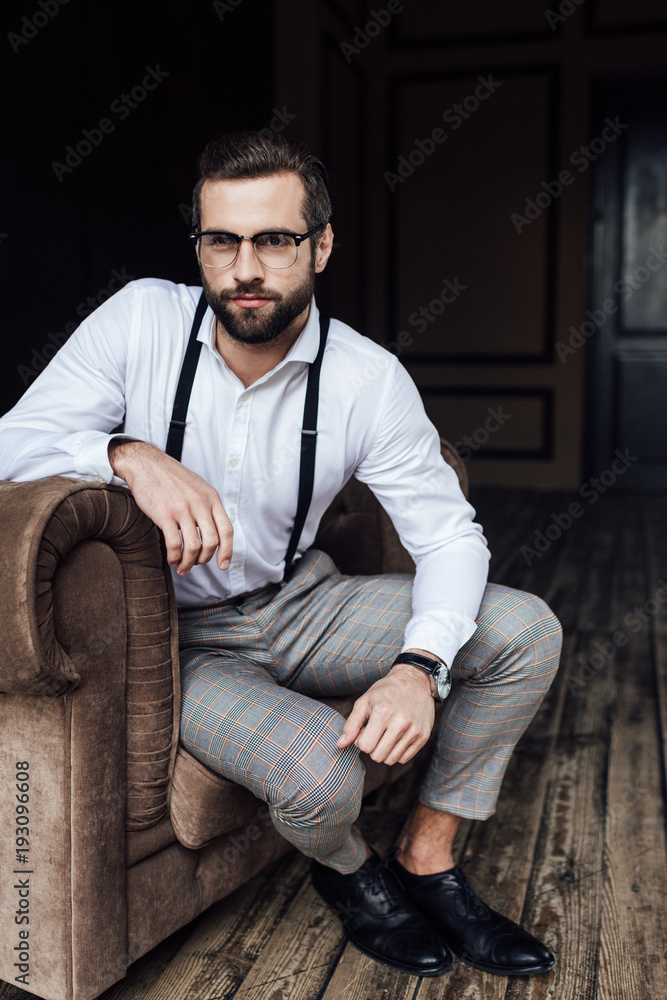handsome bearded man in eyeglasses and suspenders sitting in armchair