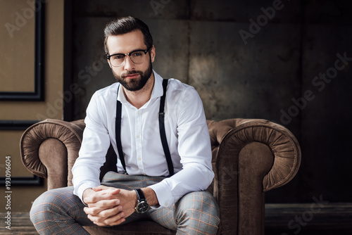 handsome stylish man in glasses and suspenders sitting in armchair