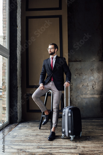 elegant businessman in suit posing on stool with luggage