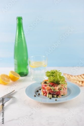 Tar-tar from salmon with avocado and tomatoes on a blue plate. Next to the plate are cutlery and toast. In the background, a lemon, a glass of water and a bottle. Light background. Vertical shot.