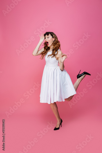 Adorable caucasian woman in white dress taking off sunglasses and looking aside on copyspace with surprise, isolated over pink background