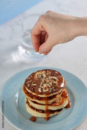 Pancakes are laid one on another, are poured with caramel sauce and sprinkled with grated almonds. Over the pancakes is brought a hand, sprinkled with pancakes nuts. Vertical shot.