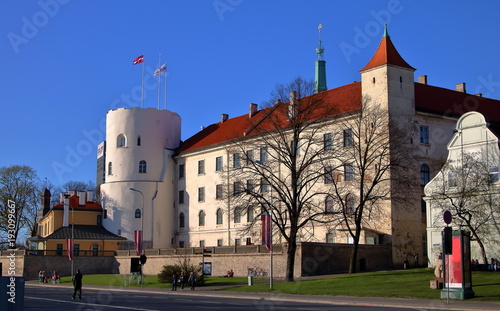 Zamek w Rydze, stolicy Łotwy, mały park przed budowlą, ludzie spacerują, siedzą na ławkach i na trawie, słoneczny dzień, błękitne bezchmurne niebo