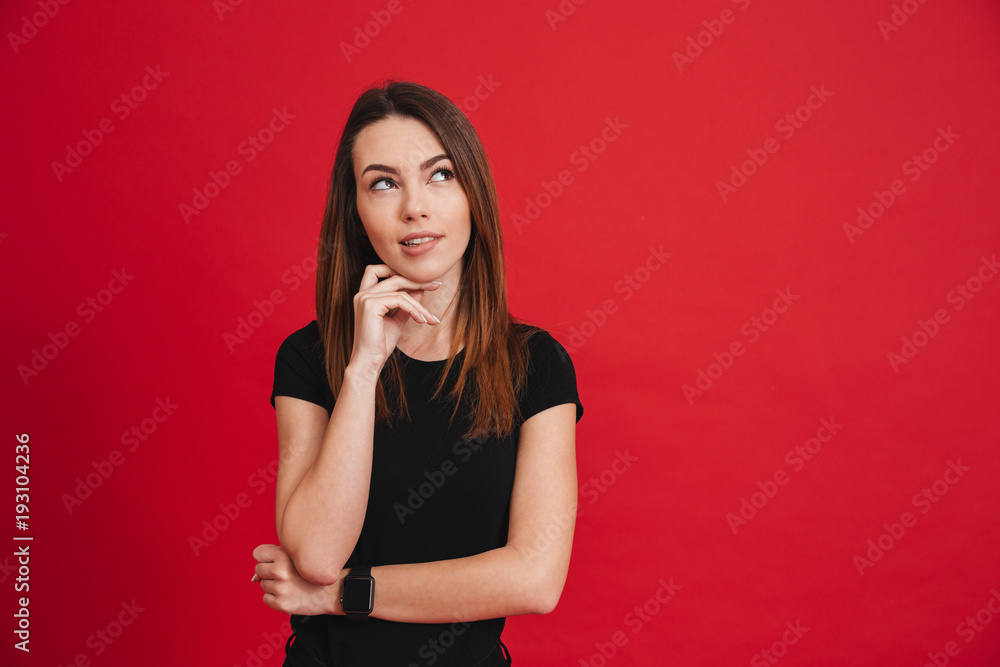Portrait of young woman 20s wearing black looking upward and touching chin with brooding or dreaming view isolated over red background Portrait of young woman 20s wearing black looking upward and touching chin with brooding or dreaming view isolated over red background