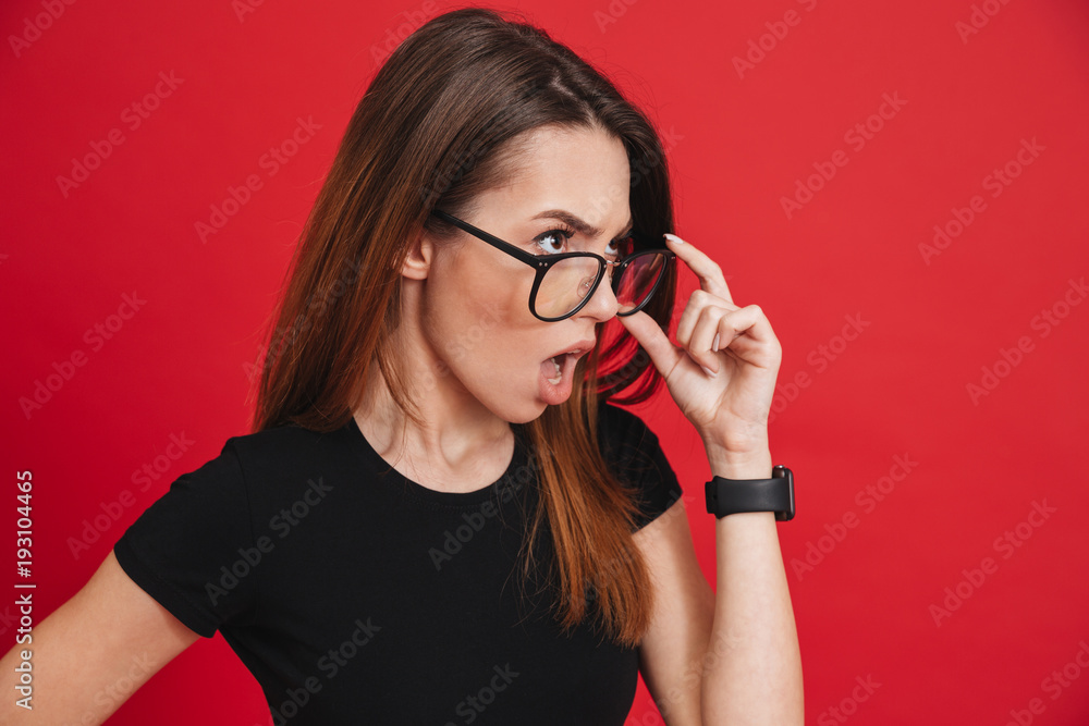 Photo of young outrageous woman wearing black t-shirt taking off glasses and looking aside with indignation or shock isolated over red background Photo of young outrageous woman wearing black t-shirt taking off glasses and looking aside with indignation or shock isolated over red background