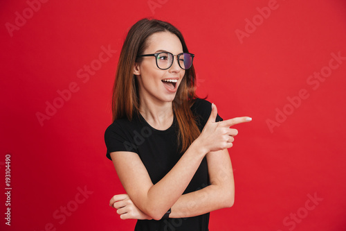 Image of cheerful woman with long brown hair winking and showing index finger aside meaning hey you, isolated over red background