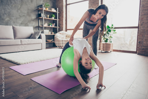 People ans sport concept. Charming small cute funny joyful active sportive girl is lying upside-down on light green fit-ball, touching floor with hands, pretty confident careful mother gives support photo