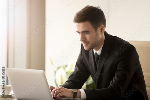 Attractive man in business suit using laptop while sitting at desk. Millennial businessman reading e-mail, surfing in Internet, communicating with colleagues online while working on computer in office