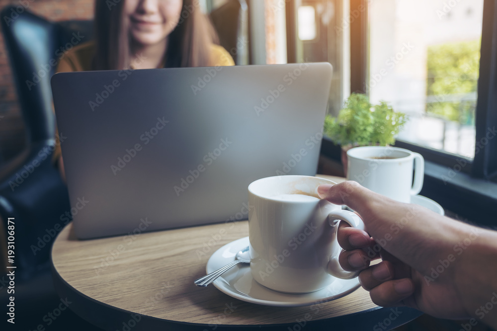 Closeup image of a hand holding a cup of coffee to drink with a beautiful Asian woman working and typing on laptop keyboard in background