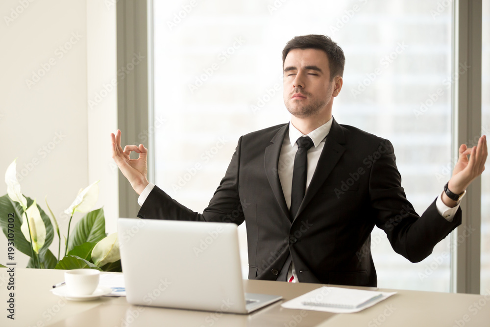 Handsome man in business suit meditating with closed eyes at work desk with laptop. Successful businessman practicing eastern spiritual practices at office workplace for relaxing and stress relief Handsome man in business suit meditating with closed eyes at work desk with laptop. Successful businessman practicing eastern spiritual practices at office workplace for relaxing and stress relief