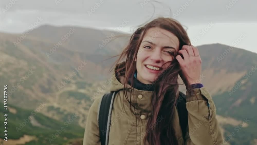 Attractive woman standing on a mountain peak with her long hair blowing in the wind smiling and trying to keep her hair off her face with her hand Attractive woman standing on a mountain peak with her long hair blowing in the wind smiling and trying to keep her hair off her face with her hand