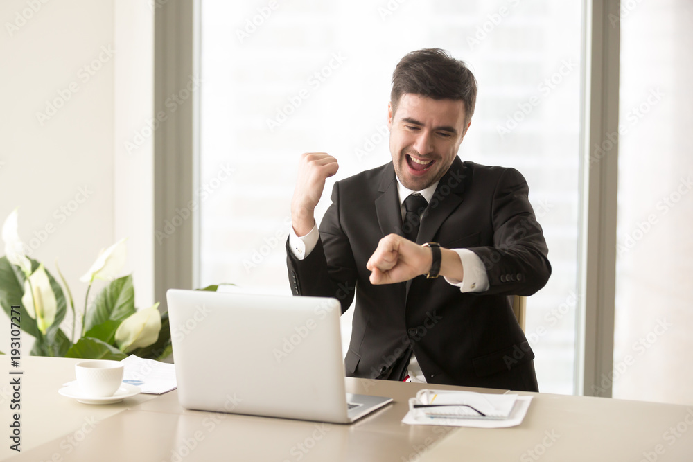Excited businessman yelling with joy when sitting at desk in front of laptop and looking at wrist watch. Happy office worker finishing work before deadline, glad because of ending of working day Excited businessman yelling with joy when sitting at desk in front of laptop and looking at wrist watch. Happy office worker finishing work before deadline, glad because of ending of working day