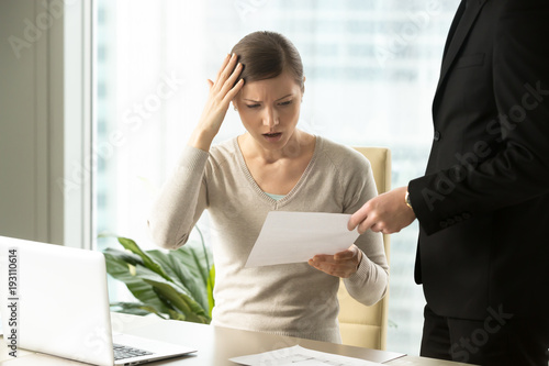 Boss giving dismissal notification to confused female office worker. Millennial woman shocked because of unexpected firing from job. Puzzled businesswoman reading lawsuit text, mistakes in document