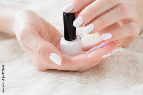 Beautiful groomed woman s hands with nail varnish bottle on the fluffy mat. Nail varnishing in white color. Manicure  pedicure beauty salon concept.