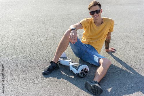 Full length portrait of cheerful teenager sitting on asphalt near gyroscope. Copy space