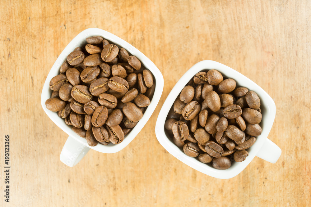 Light roast brown coffee beans in two white coffee cups on a wooden table top Light roast brown coffee beans in two white coffee cups on a wooden table top