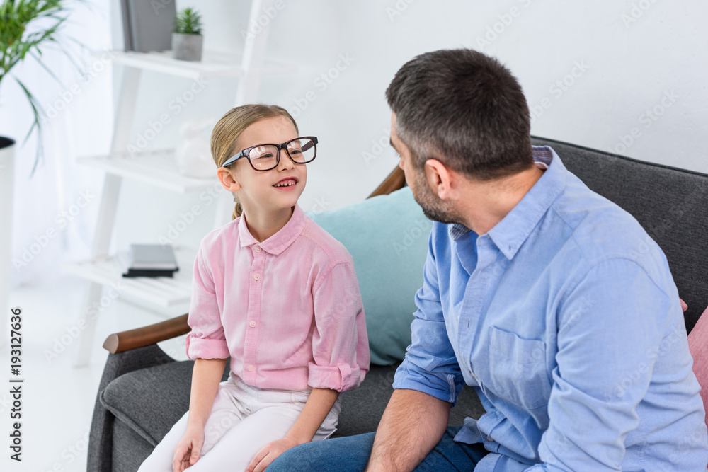 father and daughter in eyeglasses looking at each other while sitting on sofa at home father and daughter in eyeglasses looking at each other while sitting on sofa at home
