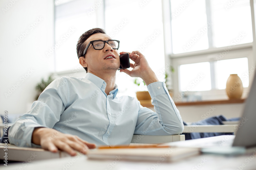 Doing business. Handsome determined dark-eyed dark-haired man talking on the phone and wearing glasses while sitting at the table Doing business. Handsome determined dark-eyed dark-haired man talking on the phone and wearing glasses while sitting at the table