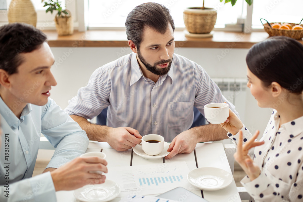 Telling news. Good-looking content bearded man drinking coffee and looking at a dark-haired woman holding a cup and talking Telling news. Good-looking content bearded man drinking coffee and looking at a dark-haired woman holding a cup and talking