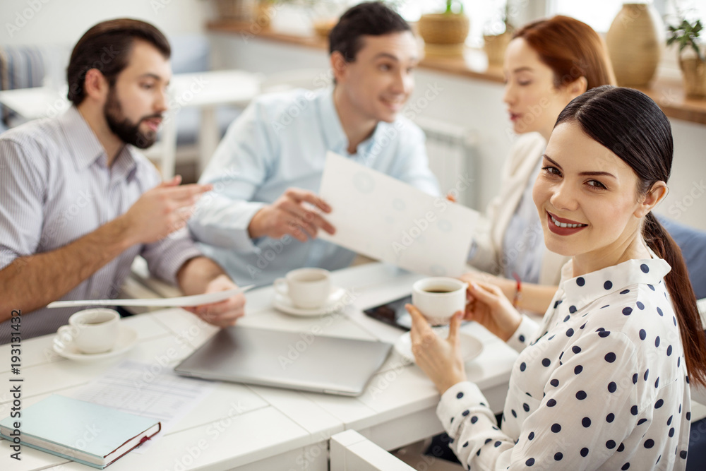 Working atmosphere. Attractive alert dark-haired young woman smiling and drinking coffee and her colleagues talking in the background Working atmosphere. Attractive alert dark-haired young woman smiling and drinking coffee and her colleagues talking in the background
