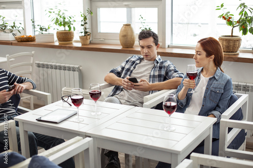 Relaxing together. Handsome concentrated dark-haired young man using his phone and his friends sitting at the table with him and drinking wine
