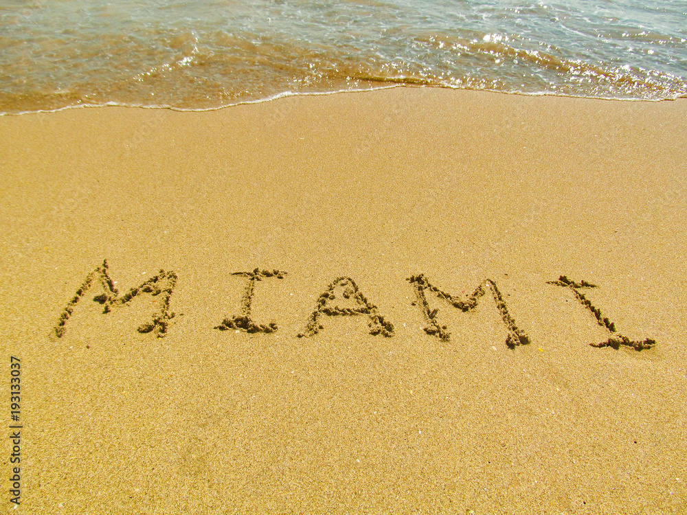 Fototapeta premium Hand drawn word on sand beach in Miami, Florida, USA. Close up detailed image of sand, text and water. Summer vacation and travel concept.