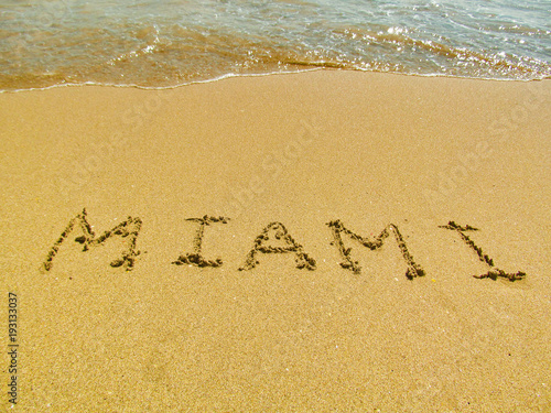 Hand drawn word on sand beach in Miami, Florida, USA. Close up detailed image of sand, text and water. Summer vacation and travel concept.