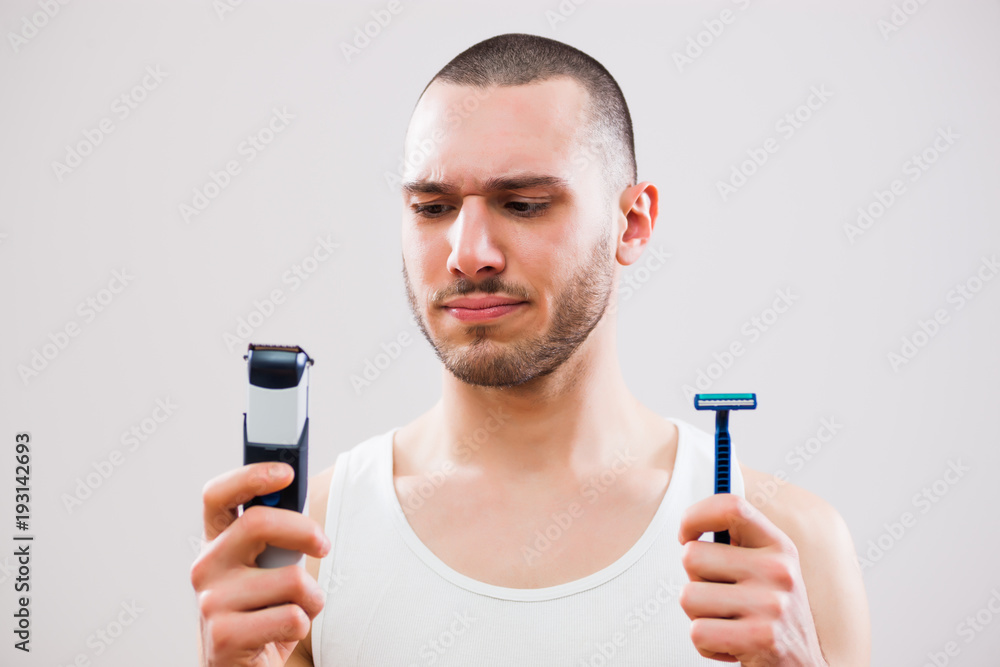Young man is holding electric and classic razor. He is deciding which one to use to shave his beard. Young man is holding electric and classic razor. He is deciding which one to use to shave his beard.