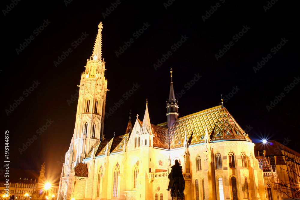 Low angle view of majestic tall Matthias Church floodlit by exterior facade lights with King Stephen equestrian statue silhouette Buda Castle Fisherman's Bastion Trinity square Budapest Hungary Europe