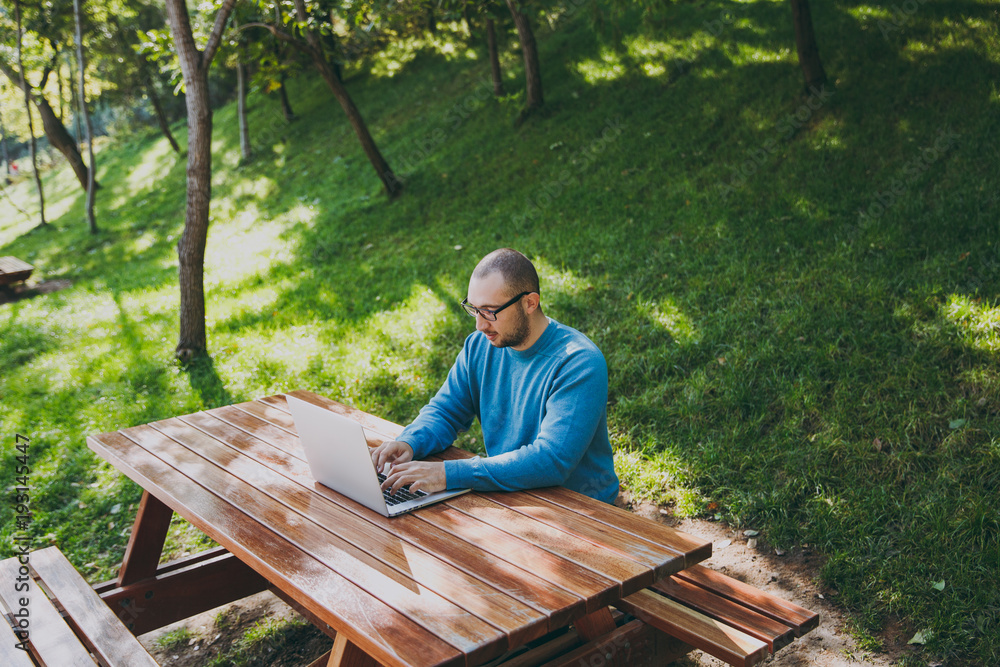 Fototapeta premium Young successful smart man businessman or student in casual blue shirt glasses sitting at table with mobile phone in city park using laptop working outdoors on green nature. Mobile Office concept.