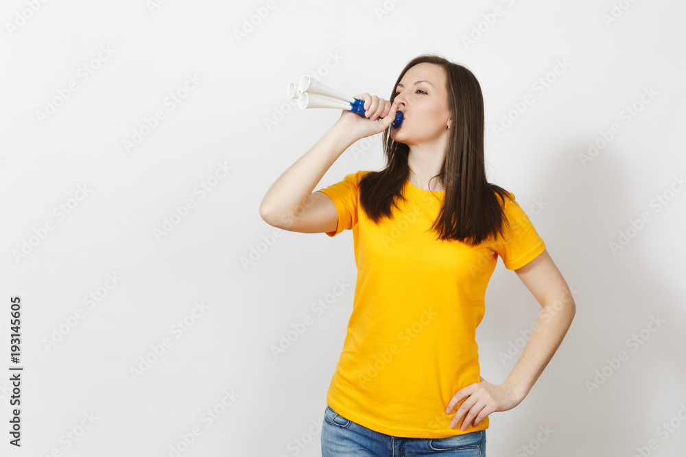 Beautiful European young happy brown-haired woman holding, blowing in football pipe, dressed in casual clothes, yellow t-shirt with shy charming smile isolated on white background. Sport fan concept. Beautiful European young happy brown-haired woman holding, blowing in football pipe, dressed in casual clothes, yellow t-shirt with shy charming smile isolated on white background. Sport fan concept.