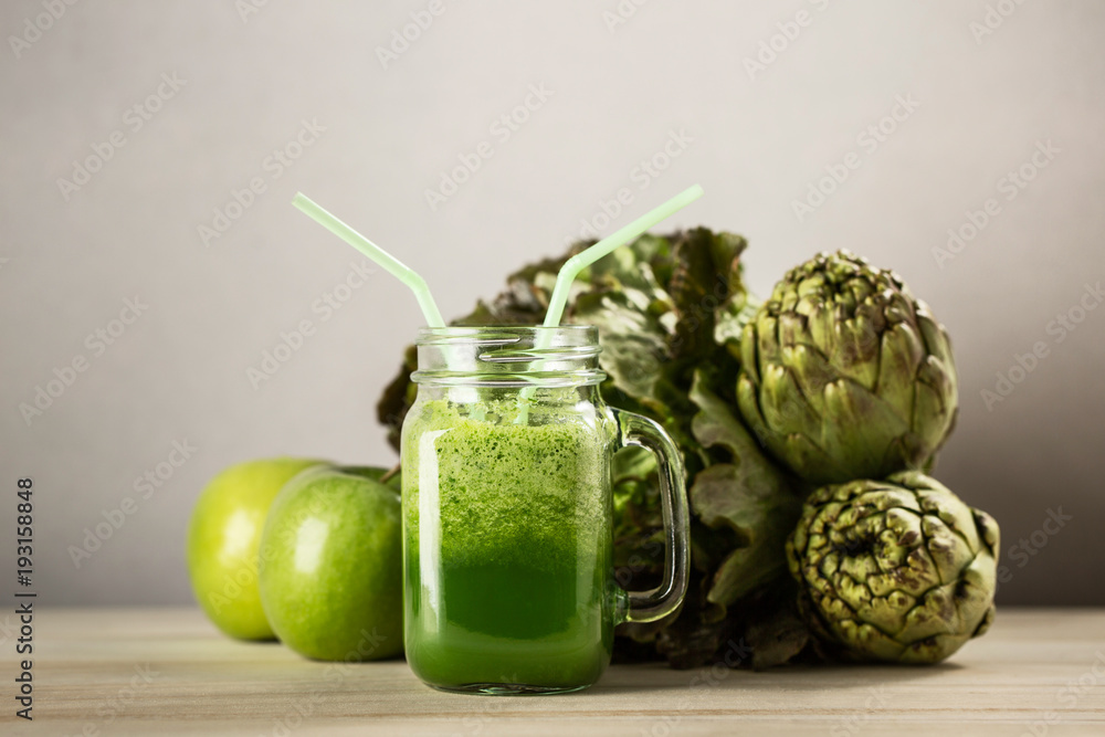 Green detox juice jar with green vegetables with two straws and vegetables in background.