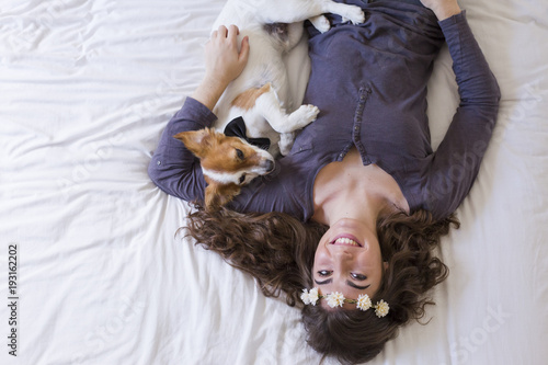 beautiful young woman lying on bed with her cute small dog besides. Home, indoors and lifestyle