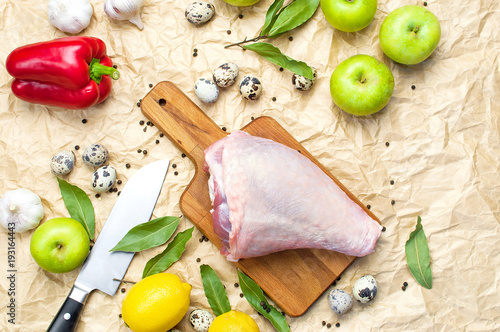 Raw turkey thigh garlic apples lemon quail eggs spices on a wooden board. Top view of a turkey, knife on a beige paper background. Culinary background, ingredients for cooking. photo