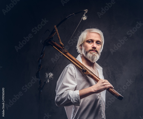 Studio portrait of a smiling handsome old man with a gray beard and white shirt holding a crossbow on shoulder.