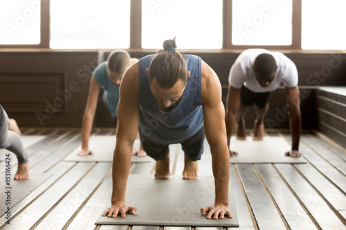 Fototapeta Naklejka Na Ścianę i Meble -  Group of young sporty people practicing yoga lesson standing in Plank pose, doing Push ups or press ups exercise, working out, indoor full length, studio