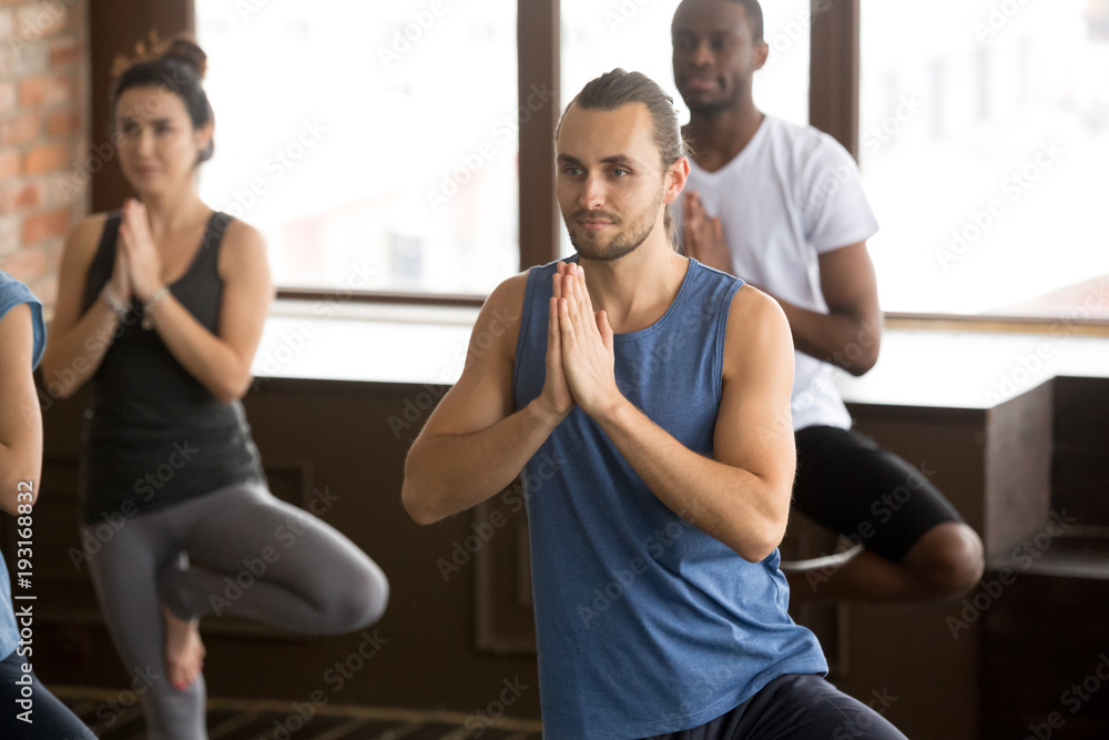 Group of young afro american and caucasian sporty people practicing yoga with instructor standing in Vrksasana exercise, Tree pose, working out, indoor close up, studio. Healthy lifestyle concept