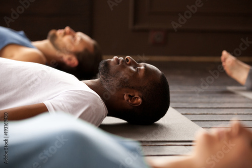 Group of young afro american and caucasian sporty people practicing yoga lesson lying in Dead Body or Corpse pose, Savasana exercise, resting after practice, working out, indoor close up, studio