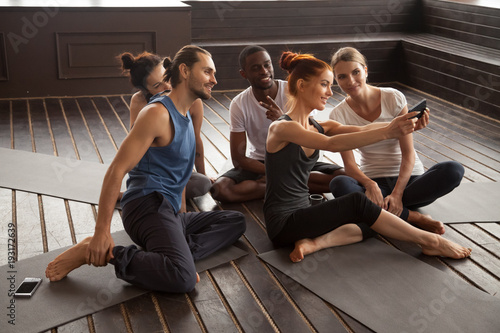Smiling young multiracial people making selfie on smartphone before yoga training sitting on mats in studio, diverse sporty fit friends taking group self-portrait photo on phone after pilates class
